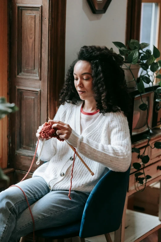 a woman sitting in a chair knitting a ball of yarn, by Julia Pishtar, pexels contest winner, curly haired, wearing a white sweater, tessa thompson inspired, white and red color scheme