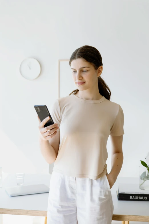 a woman standing in front of a table holding a cell phone, wear's beige shirt, silicone skin, standing in front of a mirror, relaxed posture