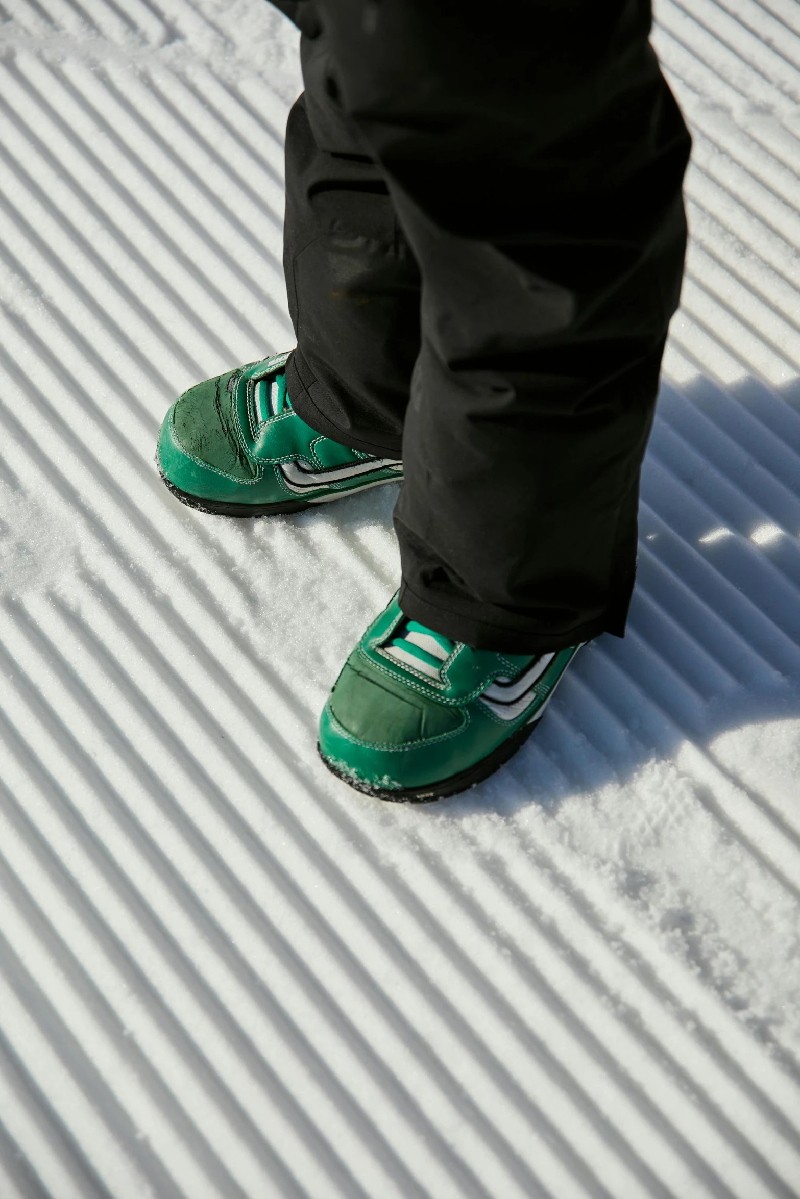 a person standing on skis in the snow, green corduroy pants, sneaker shoes, with walkways, zoomed in