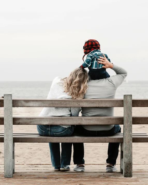 a couple of people that are sitting on a bench, by Lucia Peka, unsplash, hugging each other, on beach, non-binary, family