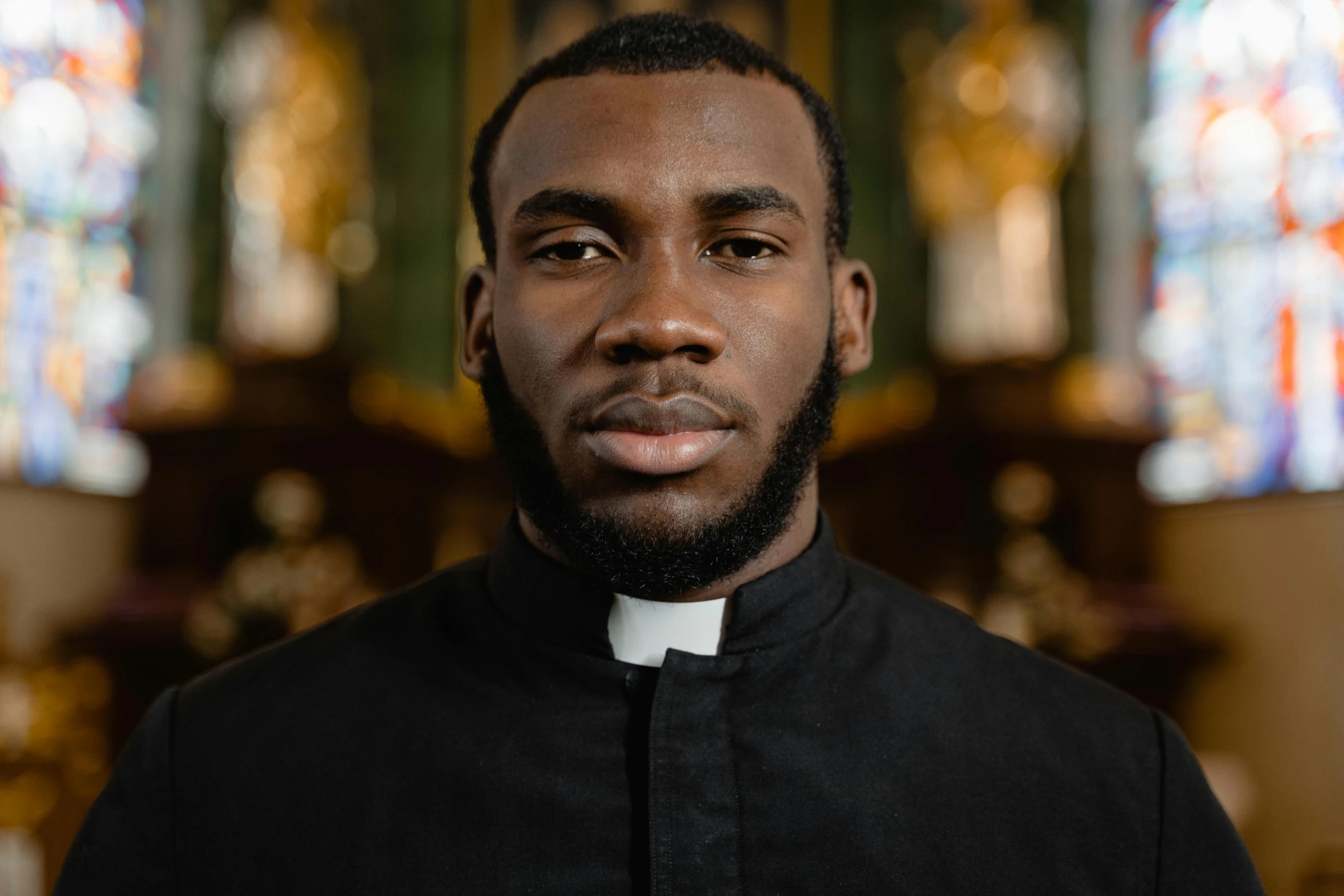 a man standing in front of a stained glass window, dark black skin tone, priest, istock, in black uniform