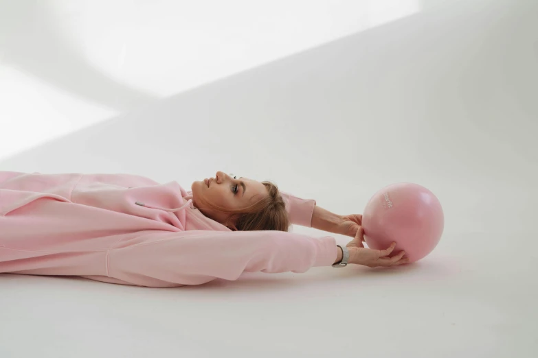 a woman laying on the ground with a pink ball, by Nina Hamnett, trending on pexels, wearing a pastel pink hoodie, on a white table, holding a balloon, head tilted down
