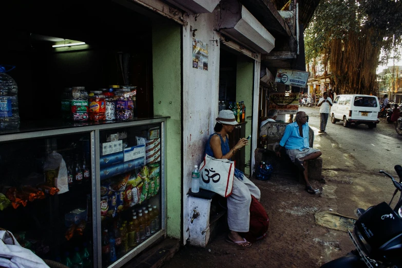 a woman sitting on a bench in front of a store, a photo, sri lanka, medium, drinking, shot on hasselblad