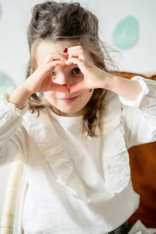 a little girl sitting at a table making a heart shape with her hands, inspired by Elsa Beskow, whiteout eyes, white, zoomed in, medium-shot