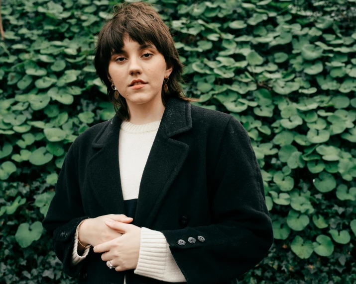 a woman standing in front of a hedge, an album cover, pexels contest winner, wearing black overcoat, rebecca sugar, charli bowater, portrait shot 8 k