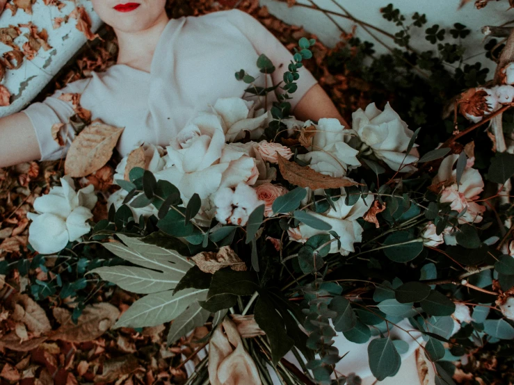 a woman laying on the ground with a bouquet of flowers, inspired by Elsa Bleda, romanticism, ivory pale skin, amongst foliage, funeral, sea - green and white clothes