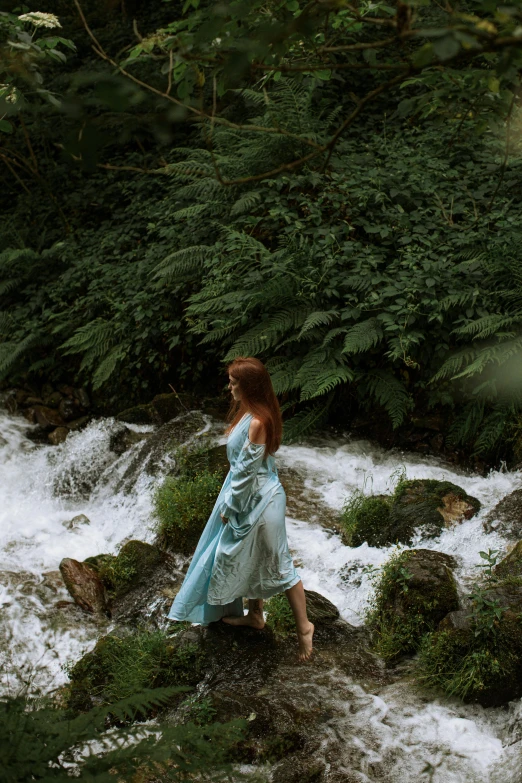 a woman in a blue dress standing on a rock next to a river, an album cover, inspired by Arthur Hughes, pexels contest winner, renaissance, walking through a lush forest, ( redhead, azores, rushing water