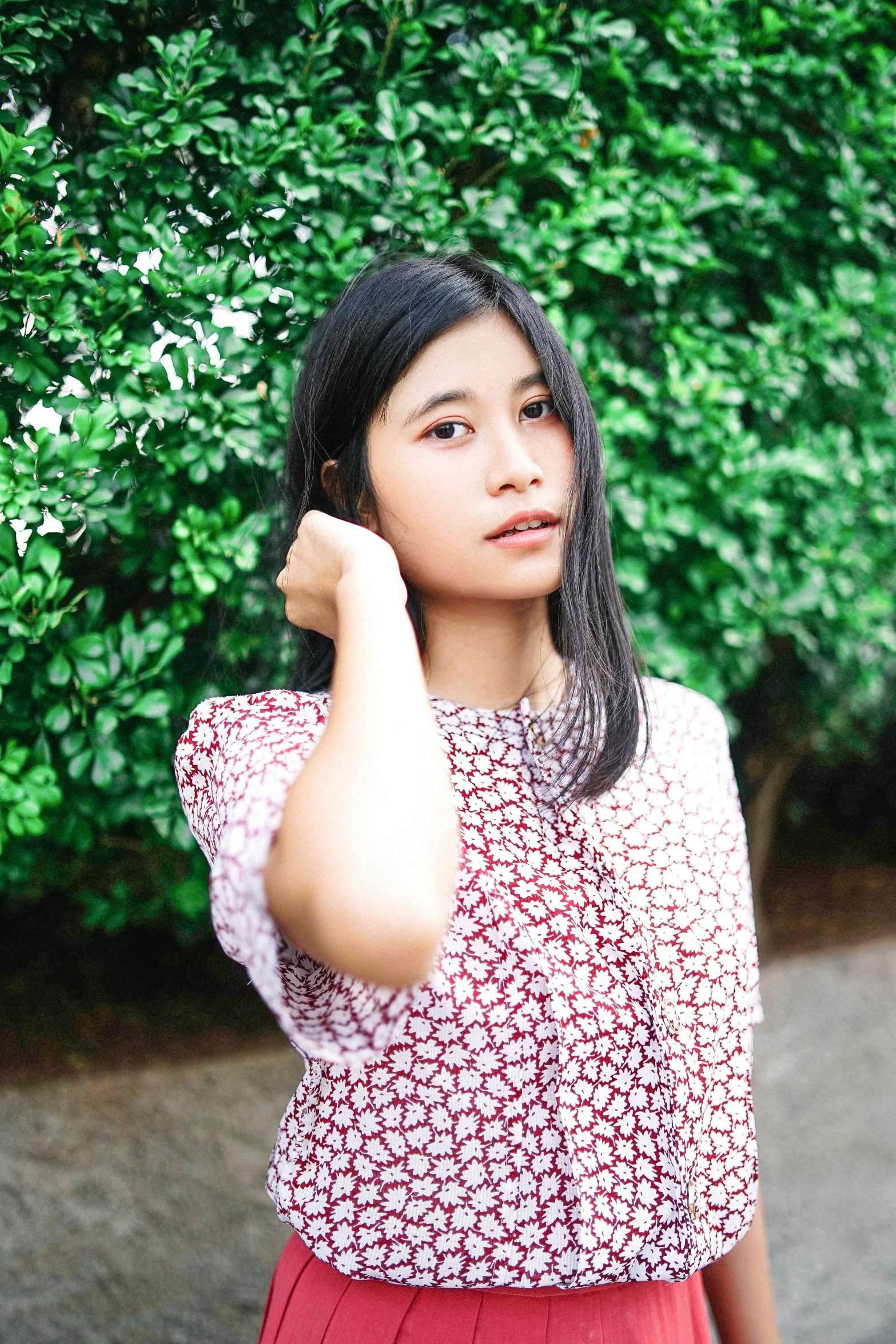 a woman standing in front of a bush with a cell phone to her ear, inspired by Ruth Jên, instagram, hurufiyya, red floral dress, profile image, wearing a light shirt, young asian woman