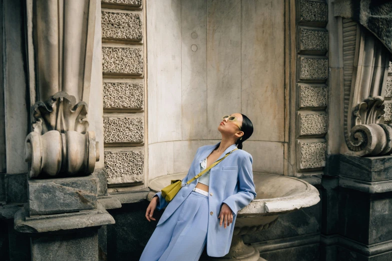 a woman in a blue coat leaning against a wall, by Anita Malfatti, pexels contest winner, wearing a light blue suit, on a marble pedestal, relaxing, doja cat