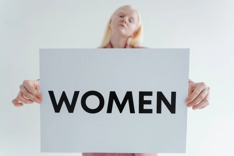 a woman holding a sign that says women, by Harriet Zeitlin, trending on unsplash, on a pale background, albino white pale skin, on grey background, blonde women
