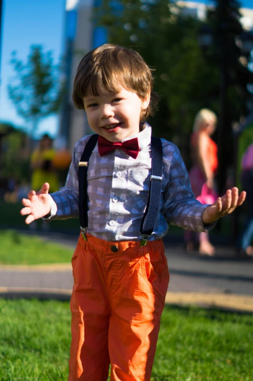 a little boy that is standing in the grass, wearing a colorful men's suit, suspenders, wearing an orange t-shirt, in a city park