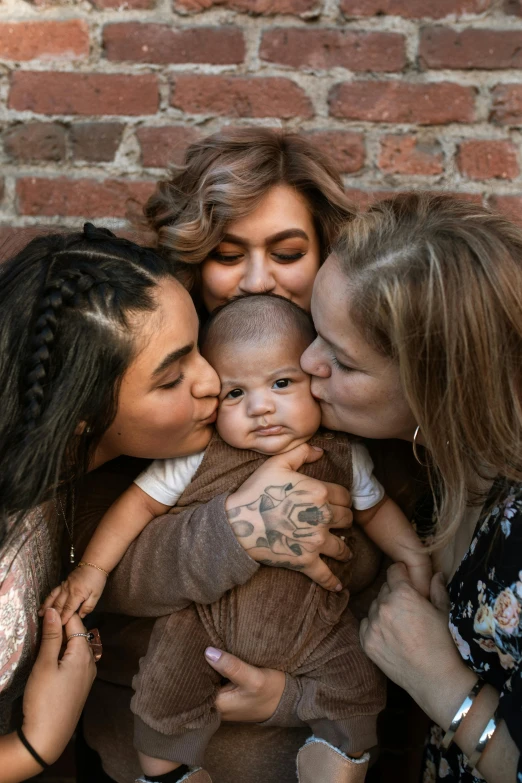 a group of women holding a baby in front of a brick wall, a picture, trending on unsplash, lesbian embrace, slide show, tattooed, square