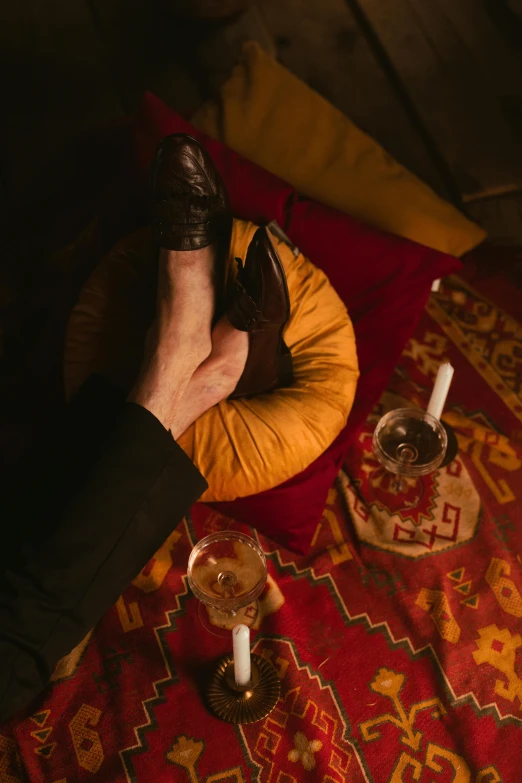 a man sitting on top of a red rug next to a glass of wine, by Elsa Bleda, renaissance, leather cuffs around wrists, arabian nights, yellow and charcoal leather, conversation pit