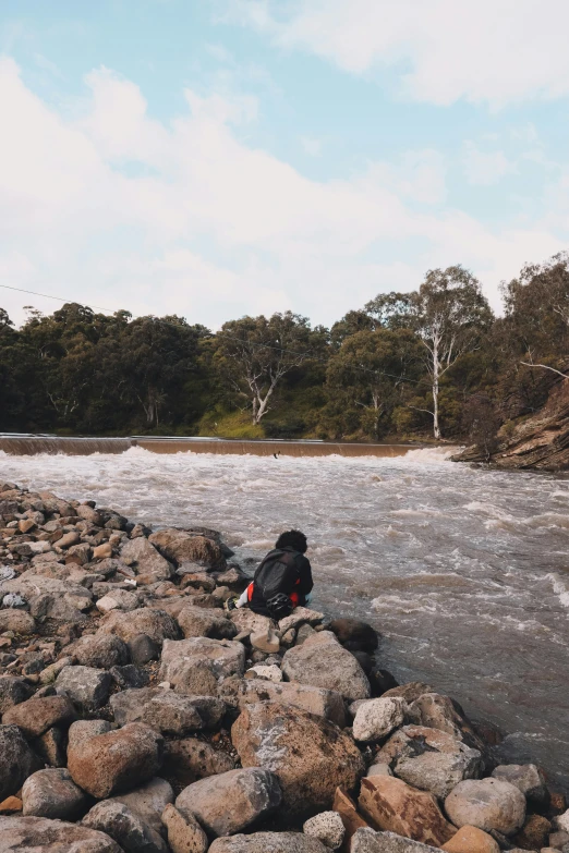 a backpack sitting on a rock next to a river, by Peter Churcher, happening, man sitting facing away, sydney park, rough water, dry river bed