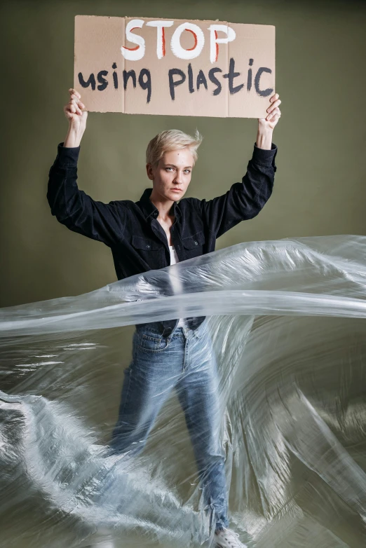 a man holding a sign that says stop using plastic, an album cover, inspired by Anna Füssli, portrait of ((charlize theron)), flowing fabric, nonbinary model, standing in a maelstrom