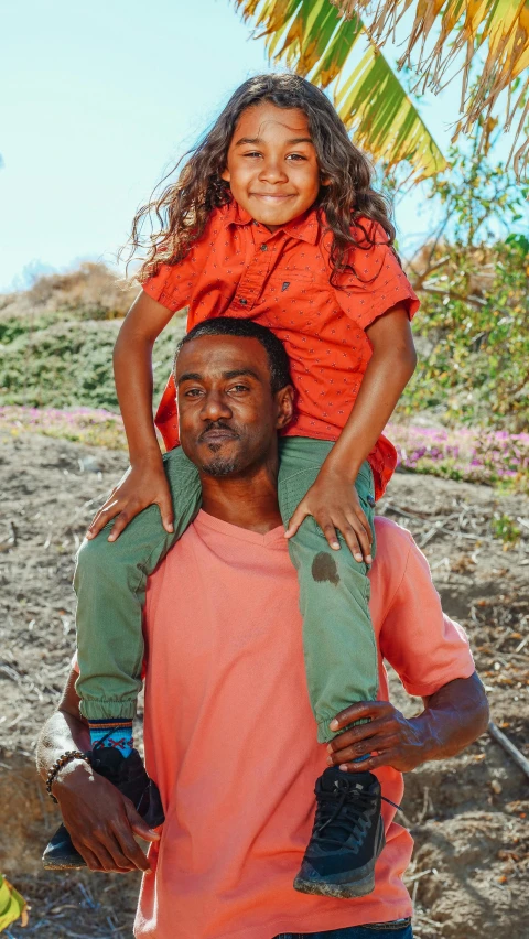 a man carrying a little girl on his shoulders, by Jessie Algie, pexels contest winner, hurufiyya, red shirt brown pants, brown skin like soil, ready to model, promo still