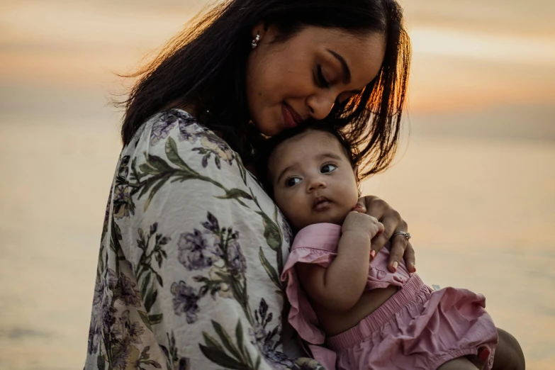 a woman holding a baby near a body of water, pink golden hour, indian girl with brown skin, profile image, manuka