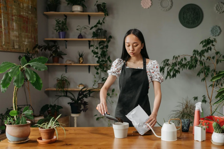 a woman pouring milk into a cup on top of a wooden table, by Julia Pishtar, pexels contest winner, lush plants and bonsai trees, avatar image, leather apron, young asian woman