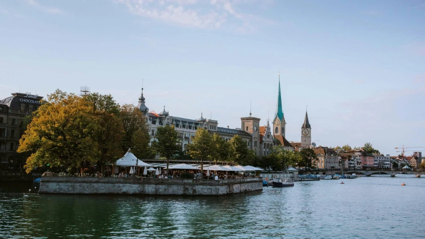 a river filled with lots of water next to tall buildings, by Julia Pishtar, photo of zurich, tall stone spires, fresh bakeries in the background, late summer evening