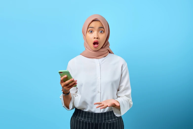 a woman with a surprised look on her face holding a cell phone, shutterstock, hurufiyya, sea - green and white clothes, malaysian, trending on tiktok, arabic pronunciation: [kaʕ.bah])