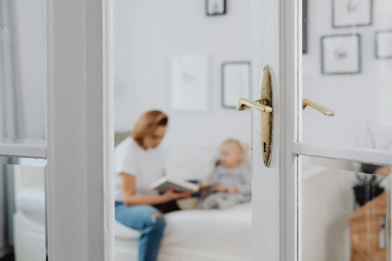 a woman sitting on a couch reading a book, by Adam Marczyński, pexels contest winner, leaning on door, with a kid, white with gold accents, childs bedroom