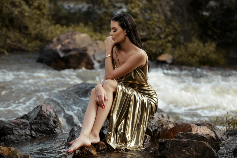 a woman sitting on a rock next to a river, an album cover, pexels contest winner, hurufiyya, elegant gold skin, selena gomez, golden dress, youtube thumbnail