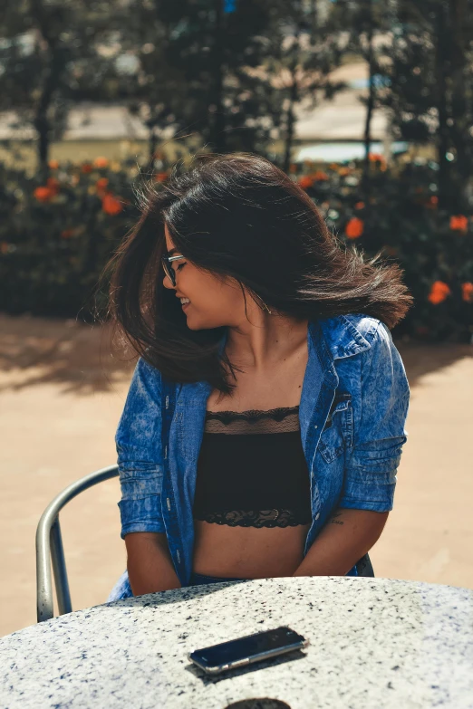 a woman sitting at a table with a cell phone, pexels contest winner, croptop and shorts, wearing a jeans jackets, avatar image, bralette