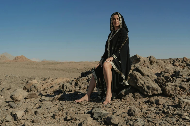 a woman sitting on top of a pile of rocks, by Alice Mason, hurufiyya, metal chain and black cape, desert robe, official store photo, arab inspired