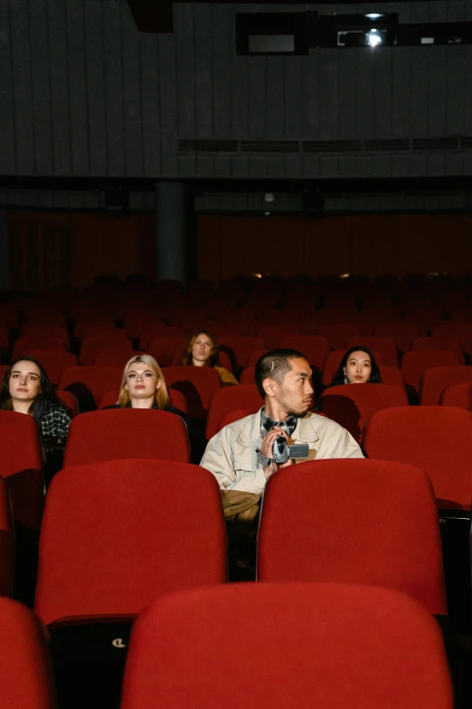 a group of people sitting in rows of red chairs, rex orange county, ( ( theatrical ) ), ignant, staring at viewer