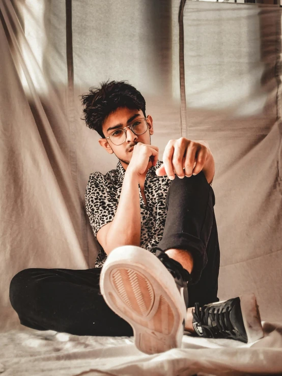 a man sitting on top of a bed next to a window, an album cover, inspired by Adam Dario Keel, trending on pexels, wears glasses, hindu aesthetic, confident stance, focus on sneakers only