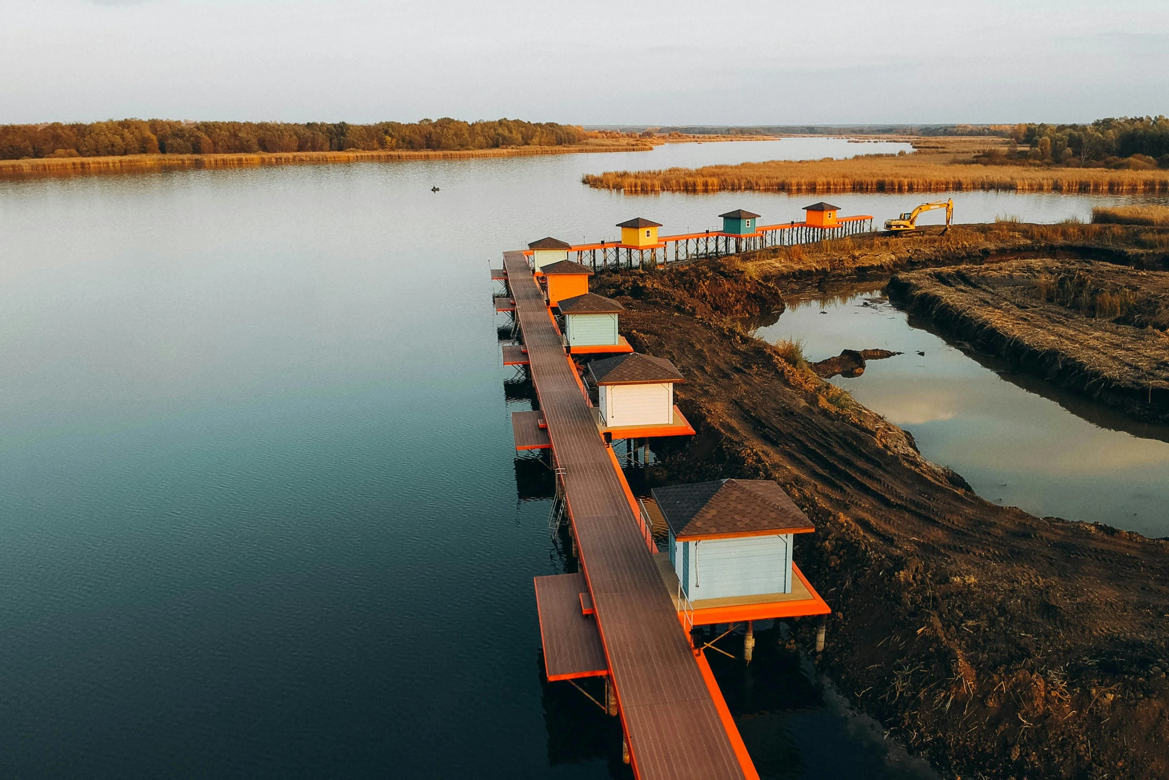 a long wooden dock next to a body of water, hurufiyya, orange roof, glamping, drone photo, huts