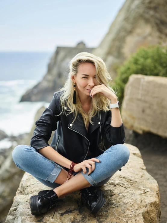 a woman sitting on top of a rock next to the ocean, a portrait, inspired by Zoë Mozert, trending on unsplash, leather jacket and denim jeans, yvonne strahovski, wearing casual clothing, sitting with wrists together