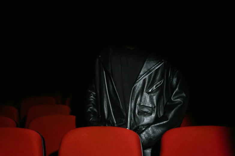 a man in a leather jacket sitting in a row of red chairs, an album cover, by Elsa Bleda, trending on pexels, realism, black jacket | shiny, in a cinema, hiding, [ theatrical ]