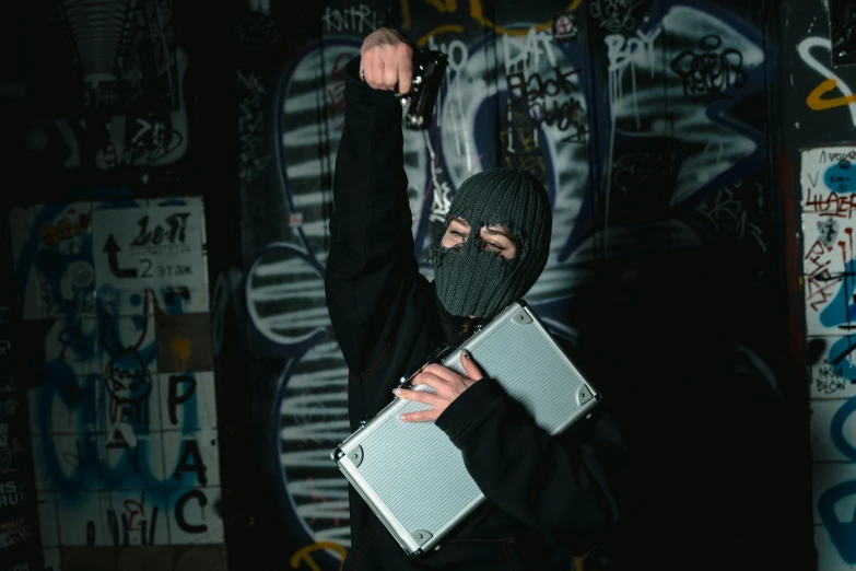 a man standing in front of a graffiti covered wall holding a briefcase, an album cover, inspired by Banksy, unsplash, wearing a dark hood, man steal computers, grey metal body, brandishing a gun