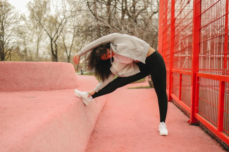 a woman stretching in front of a red fence, pexels contest winner, wearing white tights, park in background, wearing a pastel pink hoodie, a woman wearing red high heels