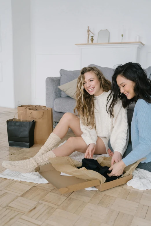 a couple of women sitting on top of a wooden floor, ecommerce photograph, smiling at each other, messy room, dressed with long fluent clothes