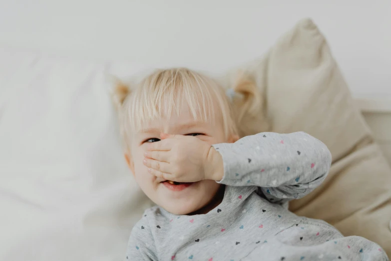 a close up of a child laying on a bed, hand over mouth, laughing, grey, large nose