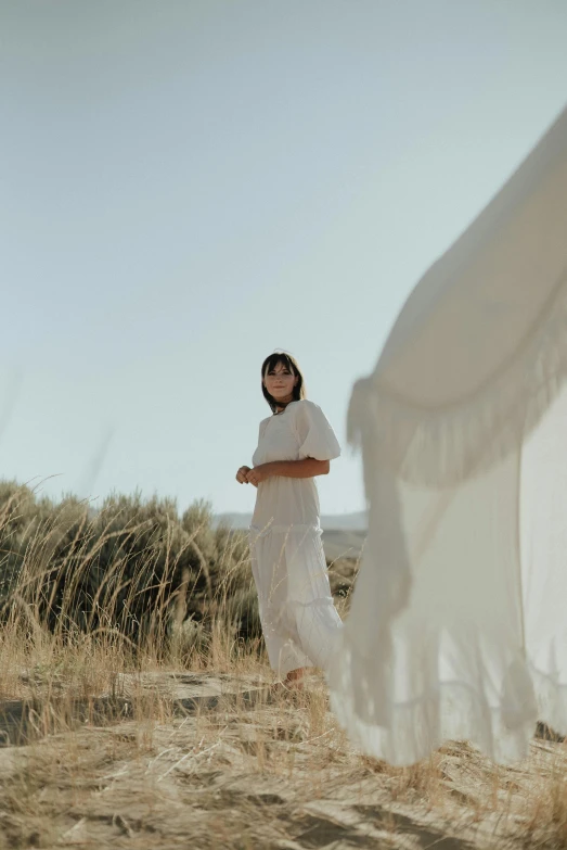 a woman in a white dress standing in a field, light and space, wearing desert poncho, in white room, glamping, slide show