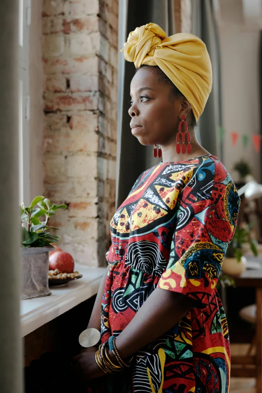 a woman with a turban standing in front of a window, a portrait, by Lily Delissa Joseph, pexels contest winner, afrofuturism, wearing red and yellow clothes, standing in a restaurant, pondering, wearing an african dress