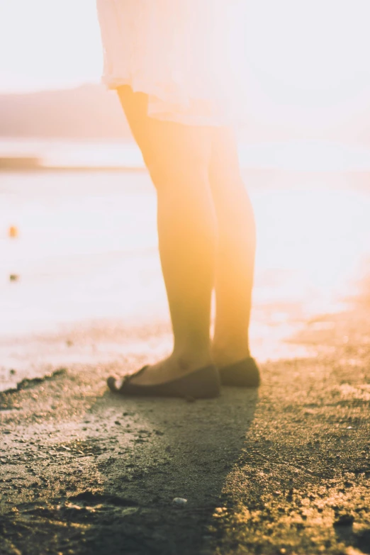 a person standing on a beach next to the ocean, soft sunlight dappling, legs intertwined, sun flares, uncropped