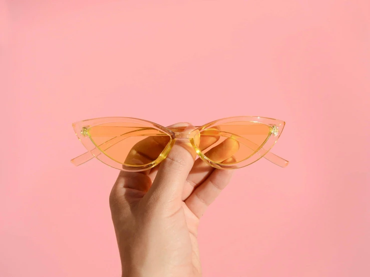 a hand holding a pair of sunglasses against a pink background, a picture, inspired by Leo Leuppi, trending on pexels, orange yellow ethereal, cat eye, translucent body, close up front view