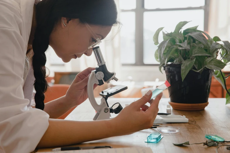 a woman in a lab coat looking through a microscope, a microscopic photo, by Liza Donnelly, trending on pexels, botanical drawing, on a wooden desk, school class, indi creates