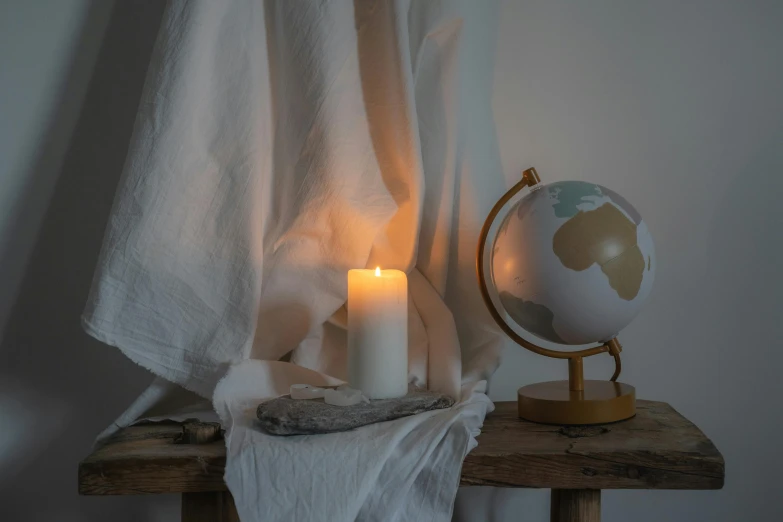 a globe sitting on top of a wooden table next to a lit candle, long flowing white robe, modern studio light soft colour, light grey, product shot