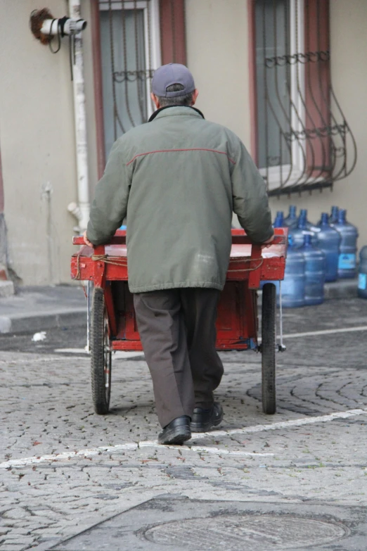 a man pushing a cart down a cobblestone street, work clothes, red boots, filling with water, facing away from camera