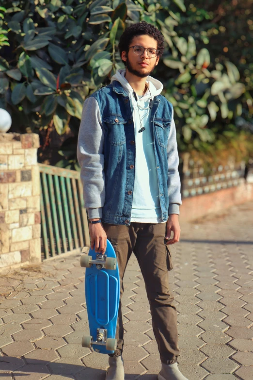 a man standing on a sidewalk holding a skateboard, a picture, inspired by Manjit Bawa, pexels, school bag, ((blue)), model is wearing techtical vest, trending on artstion