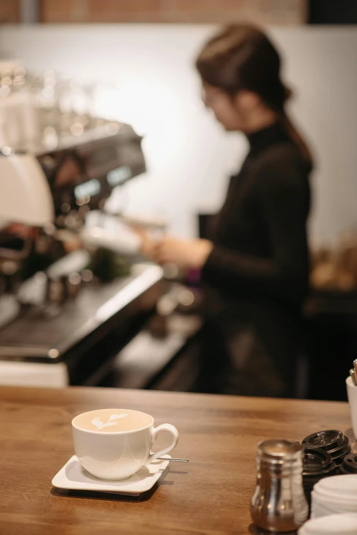 a cup of coffee sitting on top of a wooden counter, by Niko Henrichon, trending on unsplash, private press, aussie baristas, woman, coffee machine, standing in a restaurant