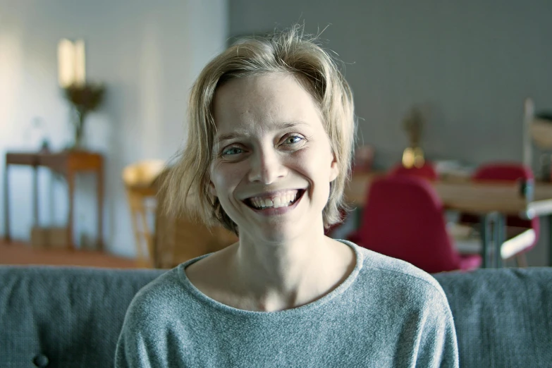 a woman sitting on a couch smiling at the camera, inspired by Louisa Matthíasdóttir, happening, close up portrait photo, animation, natural soft light, being delighted and cheerful
