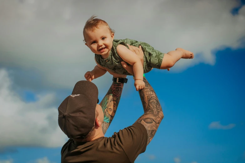 a man holding a baby up in the air, a tattoo, pexels contest winner, te pae, beautiful day, millaise and greg rutkowski, lachlan bailey