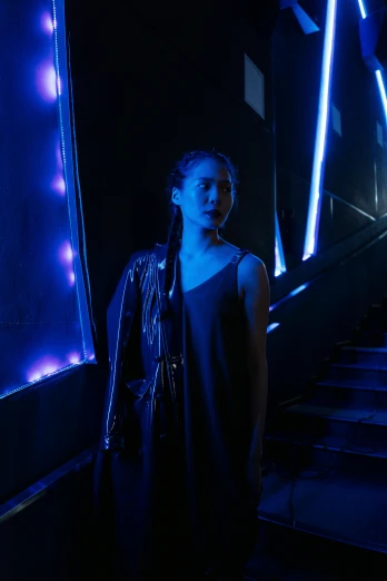 a woman standing at the bottom of a flight of stairs, inspired by Nan Goldin, neo-figurative, blue neon lighting, a young asian woman, promotional image, concert