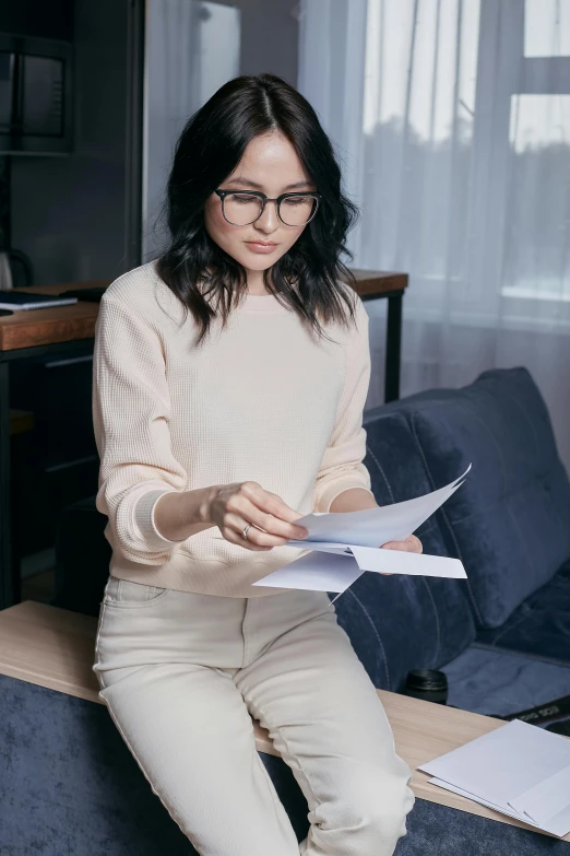 a woman sitting on a couch reading a piece of paper, tailored clothing, wearing a white sweater, rounded eyeglasses, asian descent